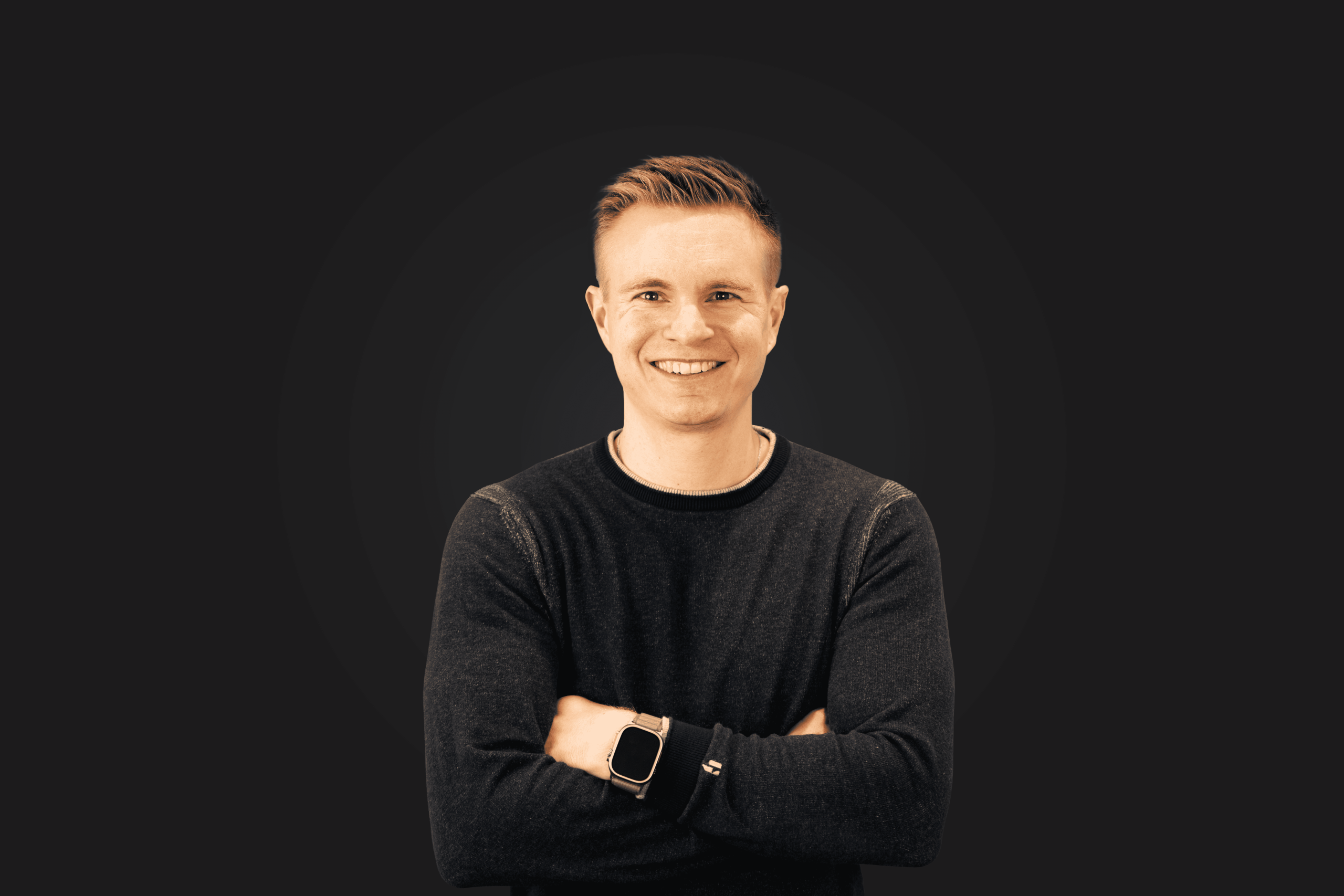 Mann mit kurzem Haar, dunklem Pullover und Smartwatch, lächelt mit verschränkten Armen vor dunklem Hintergrund.