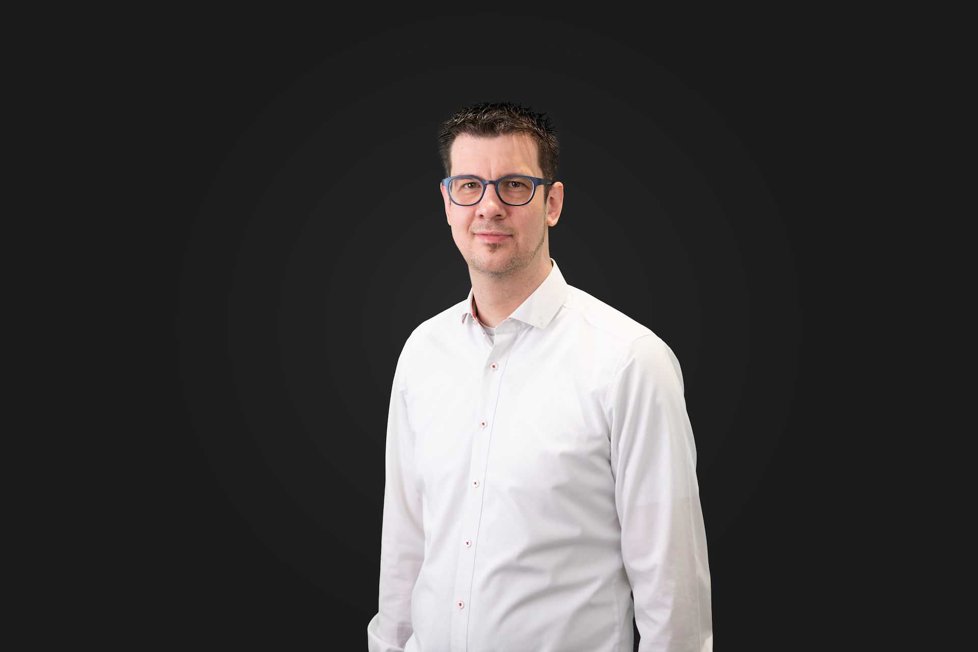Mann mit kurzen dunklen Haaren und Brille trägt ein weißes Hemd. Er steht vor dunklem Hintergrund und blickt leicht lächelnd in die Kamera.