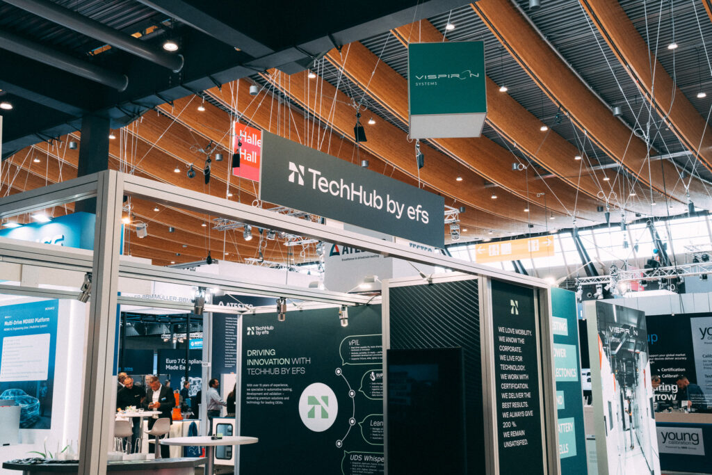 Messestand mit der Aufschrift „TechHub by efs“ auf einer Fachmesse. Der Stand ist modern gestaltet mit Infotafeln, Monitoren und Sitzbereichen. Weitere Stände und Besucher:innen sind im Hintergrund zu sehen. Die Halle hat eine hohe Decke mit Holzbalken und zahlreichen Lichtinstallationen.