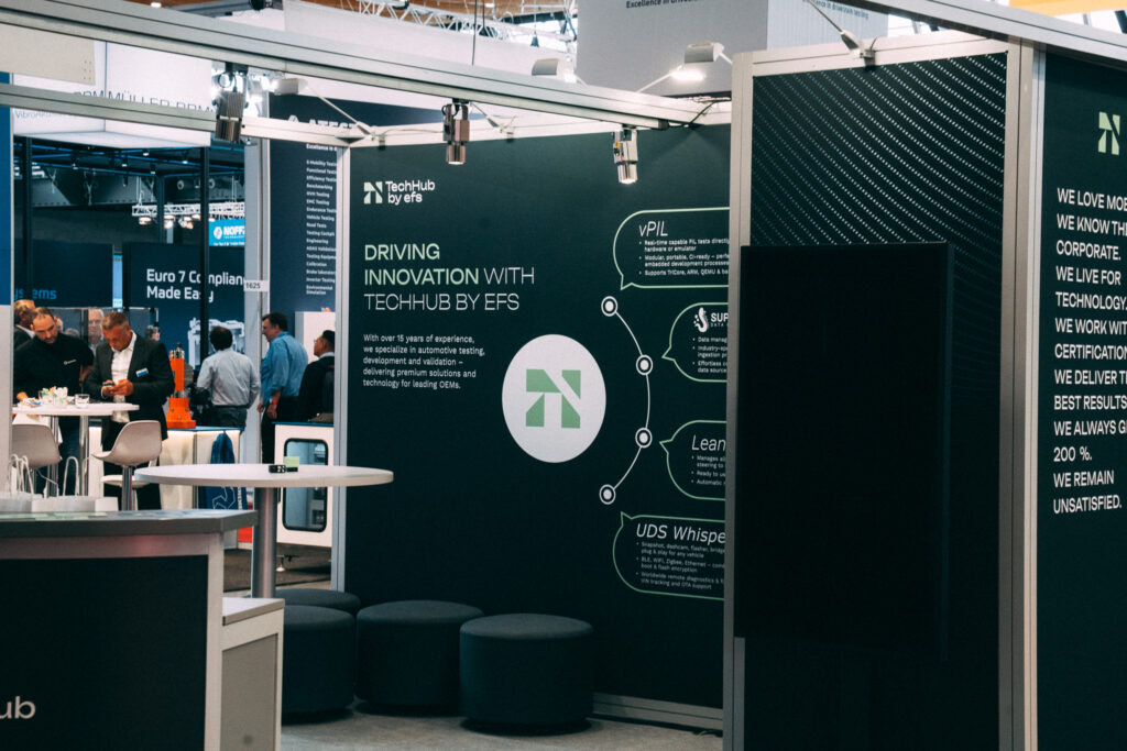 m Vordergrund ist der Messestand von EFS in Grün, Weiß und Schwarz zu sehen. In der Mitte befinden sich ein Stehtisch, eine Theke und kleine Hocker als Sitzmöglichkeit. Im Hintergrund schauen sich zahlreiche Personen an anderen Ständen um oder stehen an weiteren Hochtischen mit Stühlen.
