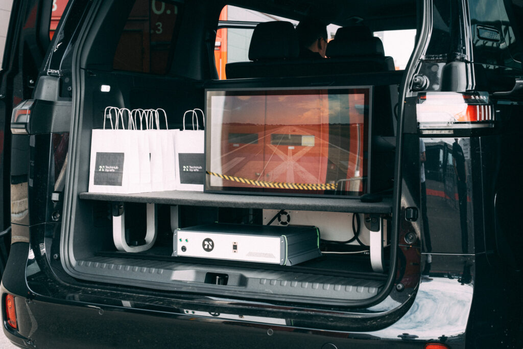 Ein großes schwarzes Auto mit geöffnetem Kofferraum, in dem mehrere weiße Tüten mit schwarzem Quadrat und weißem „Techhub by efs“-Aufdruck stehen. Im Kofferraum befinden sich zudem weitere Gegenstände. Die Fahrertür ist geöffnet, und im Inneren ist eine Person am Steuer erkennbar.