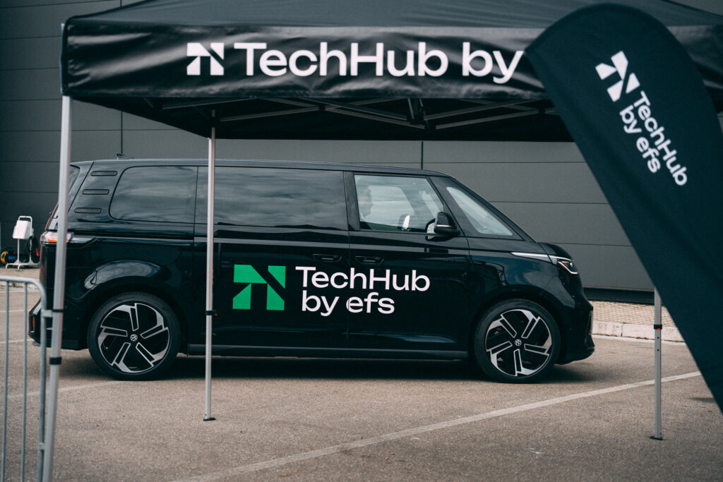 Vor einem dunklen Gebäude steht ein schwarzer VW-Bus mit dem Schriftzug ‚TechHub by efs‘. Davor sind ein Pavillon-Zelt und ein Schild platziert, beide ebenfalls mit dem Schriftzug ‚TechHub by efs‘ versehen.