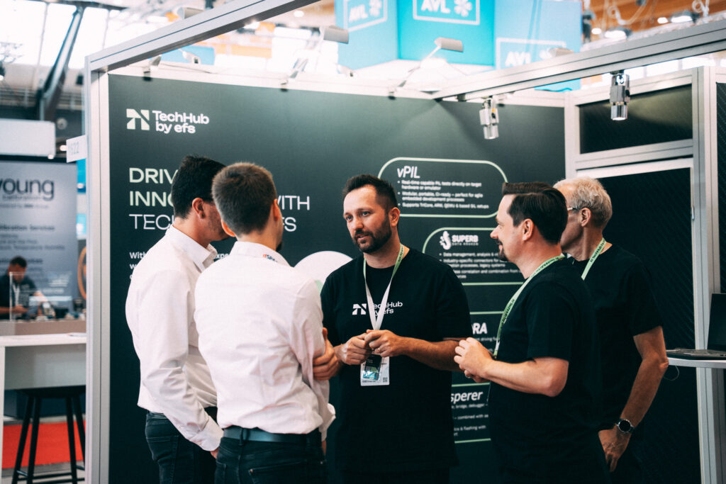 Der Messestand von Techhub by efs ist im Vordergrund zu sehen, im Hintergrund befindet sich ein weiterer Stand einer anderen Marke. Vor einer Seitenwand des efs-Stands stehen fünf Männer im Gespräch. Zwei tragen weiße Hemden und sind mit dem Rücken zur Kamera gewandt. Die anderen drei tragen schwarze T-Shirts mit weißem efs-Aufdruck sowie grüne Lanyards mit befestigten Karten.