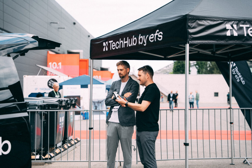 Zwei elegant gekleidete Männer stehen seitlich unter einem schwarzen Pavillon mit der Aufschrift „Techhub by efs“. Sie blicken auf ein Auto mit geöffnetem Kofferraum, das nur teilweise sichtbar ist. Im Hintergrund befinden sich eine Absperrung, ein langes schwarzes Gebäude, Mülltonnen und unscharfe Passanten.