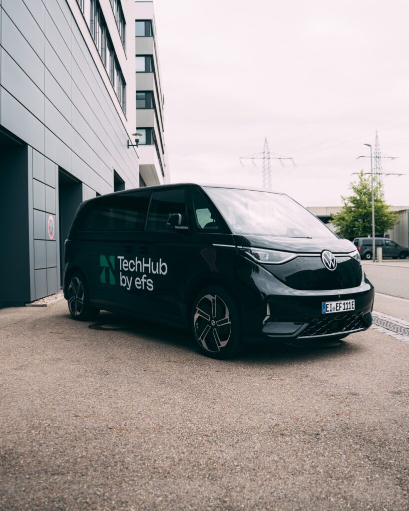 Ein schwarzer VW ID. Buzz mit dem Schriftzug „TechHub by efs“ steht vor einem modernen Bürogebäude. Auf dem Nummernschild steht „EL-EF 111E“.