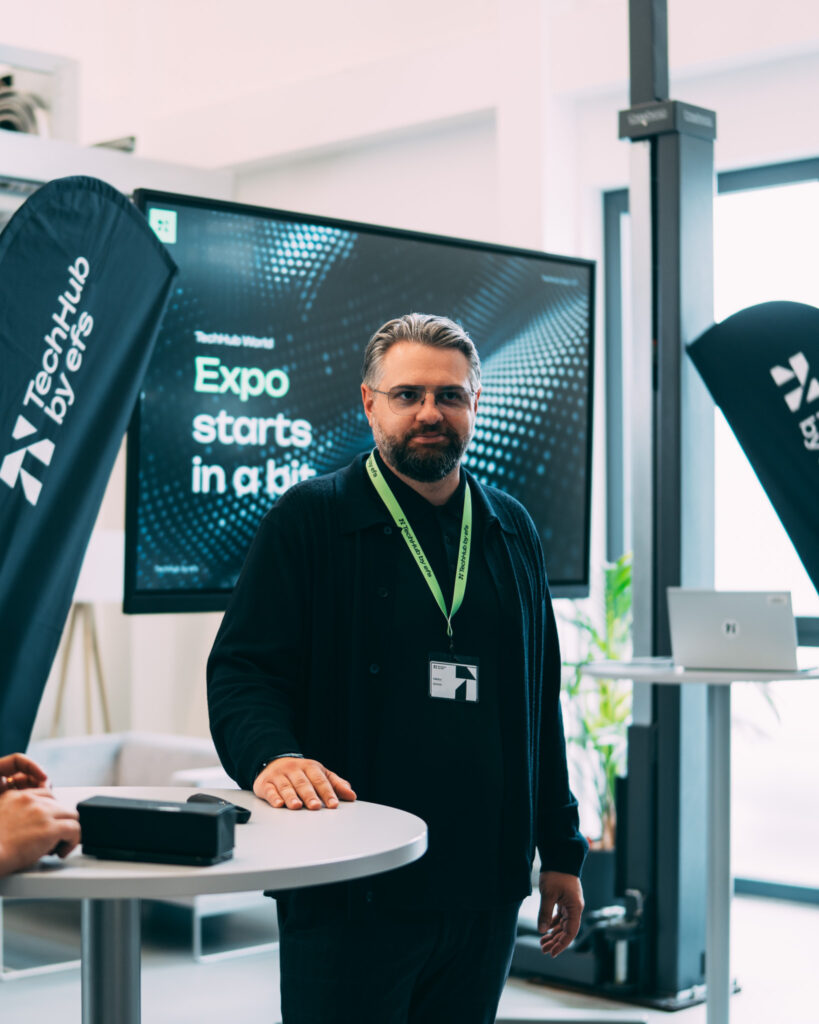 Ein Mann mit Brille und Namensschild trägt ein schwarzes Hemd und einen grünen Lanyard. Er steht vor einem Bildschirm mit der Aufschrift „Expo starts in a bit“.