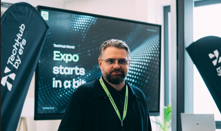 Ein Mann mit Brille und Namensschild trägt ein schwarzes Hemd und einen grünen Lanyard. Er steht vor einem Bildschirm mit der Aufschrift „Expo starts in a bit“.